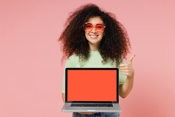 Young curly latin woman 20s wear mint t-shirt sunglasses hold laptop pc computer with blank screen mock up showing thumb up like gesture isolated on plain pastel light pink background studio portrait