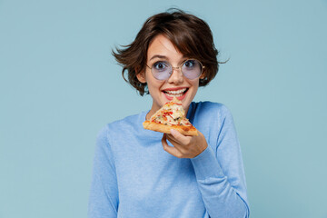 Young smiling excited caucasian woman 20s in casual sweater look camera biting eat italian slice of pizza isolated on plain pastel light blue background studio portrait. People lifestyle food concept.