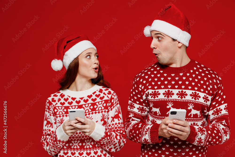 Poster Young shocked couple friends two man woman in sweater hat hold in hand use mobile cell phone look to each other isolated on plain red background Happy New Year 2022 celebration merry ho x-mas concept