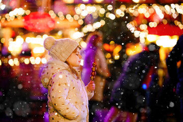Little preschool girl, cute child eating fruits, strawberries covered with chocolate with decoration and lights on background. Happy child on Christmas market in Germany.