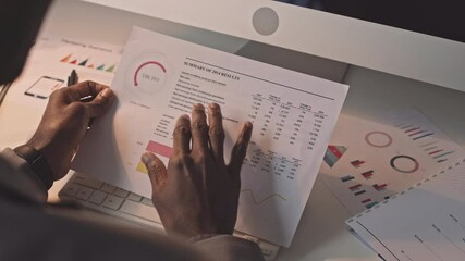 Over shoulder of businessman sitting at desk in office in evening, looking at company performance results and typing on keyboard of computer, working
