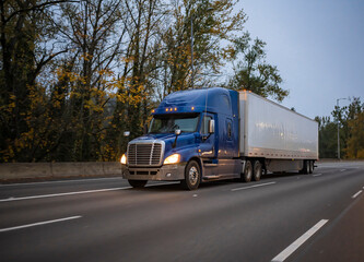Dark blue big rig semi truck with turned on headlights transporting cargo in dry van semi trailer...