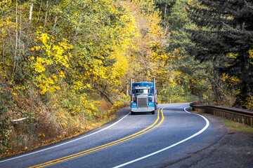 Landscape with blue big rig semi truck and bulk semi trailer running on the winding road with...