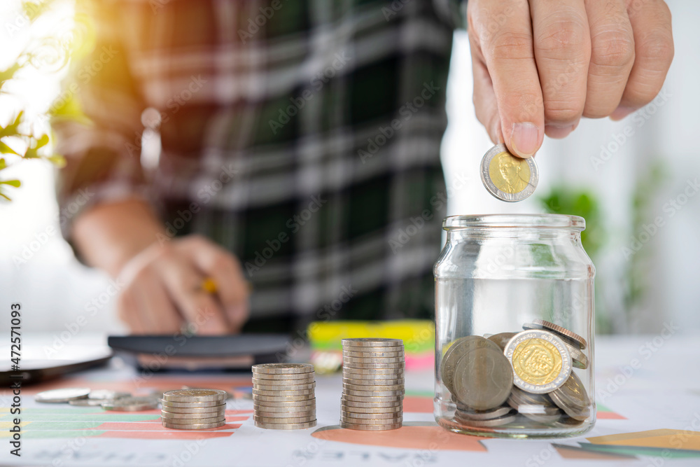 Poster closeup image of a businessman calculating, stacking and putting coins in a glass jar