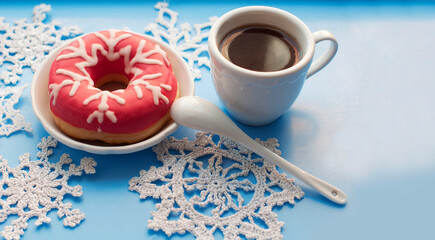 Donut with pink icing and white icing snowflake with a cup of coffee and knitted snowflakes on a blue background. Space for text