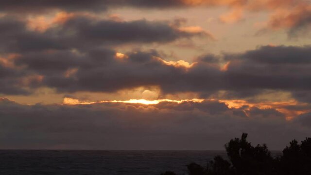 Sun approaches horizon over Indian Ocean behind grey clouds adorned with gold and orange hues. Time lapse