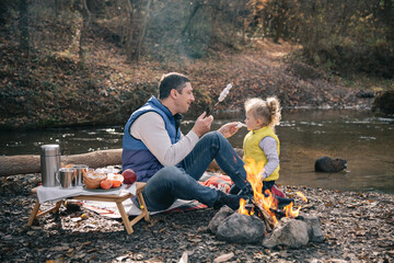 Dad and daughter fries marshmallows on sticks by the fire.