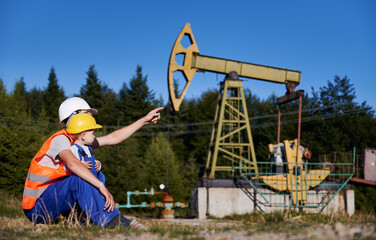 Oil rig operator introducing his son to his profession. Oilman squatting on lawn with male child, not far from the petroleum well and pointing finger to pump jack.