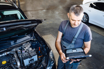Auto service, repair, maintenance concept. Skilled mechanic using a laptop computer to check collect information during work a car engine.