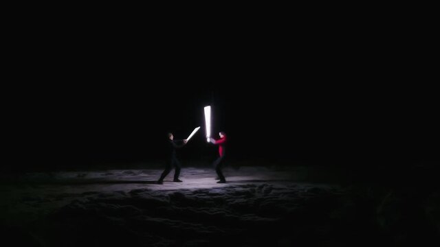 Two guys are fighting with lightsabers at night in winter in the field outdoors. Father plays with son. Friends fooling around. Funny situation. Male friendship. Raising a child. Holidays