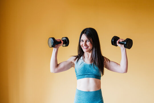 Cheerful woman exercising with dumbbell in front of yellow wall at home