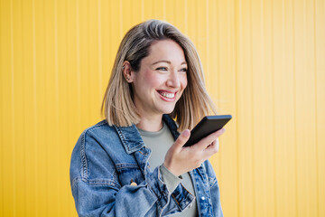 Happy woman sending voicemail through smart phone by yellow wall