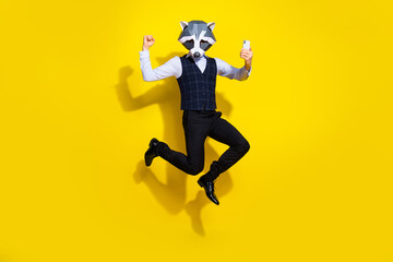 Fototapeta na wymiar Full length photo of weird guy racoon mask jump raise fist up use device wear stylish trend vest isolated over yellow color background