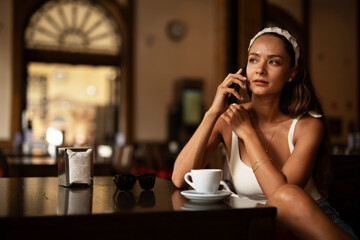 Stylish young woman drinking coffee at cafe. Beautiful girl talking to the phone while enjoying in fresh coffee