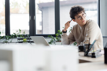 Thoughtful businessman holding pen near laptop in office.