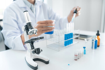 Scientist woman holding beaker and dropper to dropping blue liquid into test tube while working with microscope