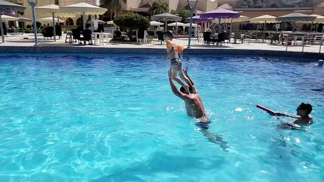 A boy-child dives in the pool water, he jumps from his father's shoulders creating a lot of splashes of water, family holidays, summer, vacation, pool, slow motion
