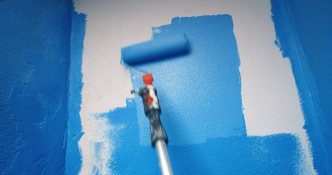 Close up shot of blue paint applied on the white wall with roller working tool. Concept of home repairs and renovation. Young woman hands with orange roller painting wall