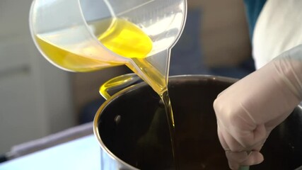 Pouring oil into a steel pot for creating a soap building mixture then stirred by hand, Close up handheld shot