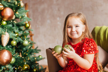 Fototapeta na wymiar Little girl in a red dress with a Christmas tree toy in her hands sits at the Christmas tree. Christmas Holidays. Christmas decor, atmosphere, winter background