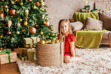 Obraz na płótnie Canvas Little girl in a red dress with a Christmas tree toy in her hands sits at the Christmas tree. Christmas Holidays. Christmas decor, atmosphere, winter background