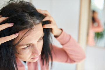 Fototapeta premium Portrait of a beautiful young woman examining her scalp and hair in front of the mirror,