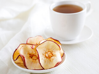 Dried thin slices, apple fruit chips on white plate and cup of tea. Healthy food and snacks. Fruits are bright orange in color. Side view close up, Light background. Minimalism