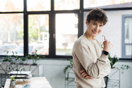 Young Transgender Person In Earphone Holding Eyeglasses And Smiling At Camera In Office.