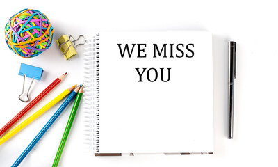 Notebook ,pencils,pen and rubber band with text WE MISS YOU on the white background