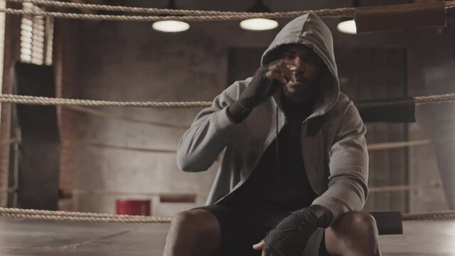 Slowmo portrait of young African-American male boxer with wrapped hands in grey zip up hoodie posing for camera sitting on boxing ring