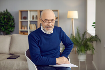 Bald bearded senior man in glasses sitting in chair at home, holding notebook on his lap and...
