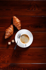 Croissants with a cup of coffee and two lumps of brown sugar lie on a wooden table. Vertical framing. View from above. Place for your text.