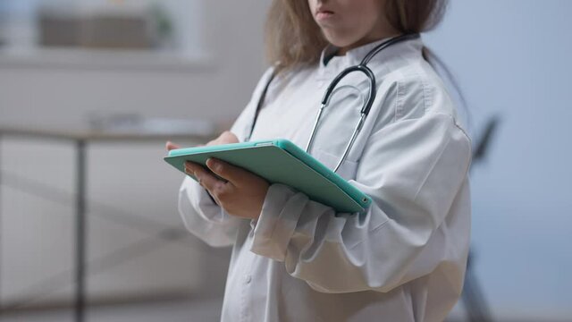 Unrecognizable little person in medical gown surfing Internet on tablet standing indoors in hospital. Young Caucasian woman thinking reading latest news online. Medicine and modern technologies