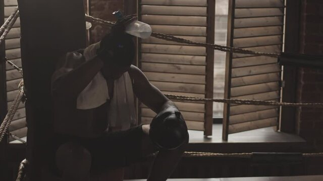 Medium slowmo shot of Black male boxer pouring water from bottle on head and body sitting in corner of boxing ring after fight