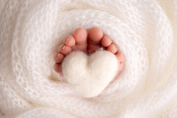 The tiny foot of a newborn baby. Soft feet of a new born in a white wool blanket. Close up of toes, heels and feet of a newborn. Knitted white heart in the legs of a baby. Studio macro photography.