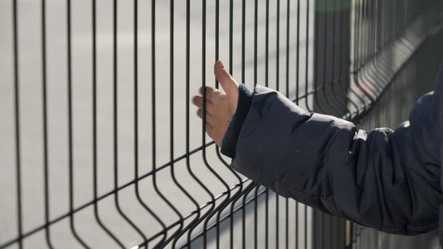 Closeup of little boy walking by the metal fence and touching it with hand. Concept of poverty, immigration, bullying and kids stress