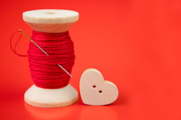 A spool of red thread, a needle and a heart button on a red background.The concept of decor, sewing,needlework.Valentine's day