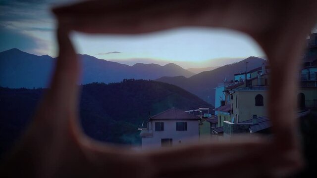 Finger Frame. Woman folding her hands in frame with a setting sun illuminates the roofs of a medieval town in the mountains. Perinaldo, Italy.