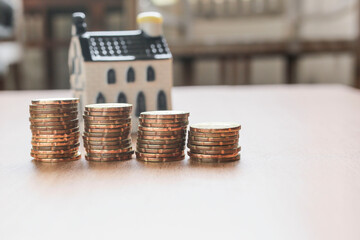 Coins are stacked on the wooden table with a house model at the back. Business and financial concept. Noise is visible due to the texture of the subjects