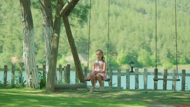 Little girl swings alone on a swing in the park by the lake.View of unhappy girl swinging on swing in park by lake at sunset.