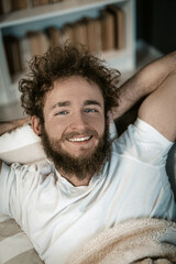 Handsome Guy with Blue Eyes, Curly Hair, a Beard and a Wide Smile Lies Under the Cover in his Bed. Top View. Close-up Portrait. Book Shelves Background