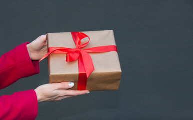 Hands of woman dressed in crimson jacket are holding a gift box wrapped in kraft paper and decorated with a red ribbon with a bow. Isolated on gray background. Outdoor