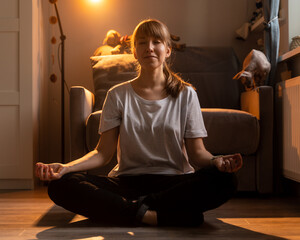 Young smiling female meditating in lotus pose. Doing yoga at home in cozy evening