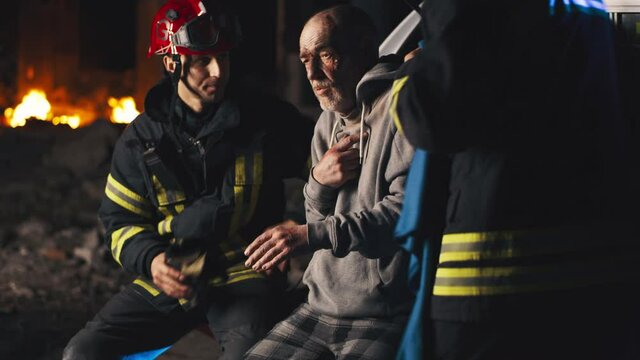 Male rescuers talking and putting blanket on shoulders of elderly man while paramedic wrapping bandage around injured hand of patient during work on disaster site at night