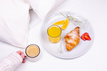 Romantic breakfast in bed. Woman hand with mug of coffee, croissant, strawberry, orange juice on the ceramic plate. flat lay top view image.