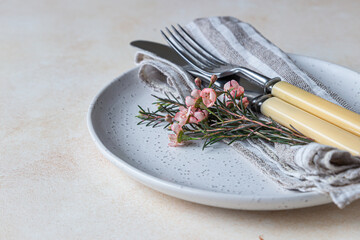 Romantic or spring table setting. Knife and fork, little pink flowers and linen napkin on a plate, light concrete background.