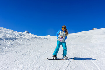 Fototapeta na wymiar A girl snowboarder in ski goggles with loose hair stands alone on a ski slope. In the background snowy mountains and clear blue sky. Active winter holidays in the mountains.