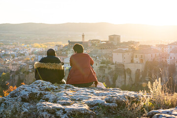 Tourist couple sitting on a rock at sunset looking at the landscape in a tourist city. Cuenca city, Spain. Concept of friendship and travel as a couple