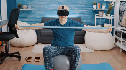 Aged person pulling resistance band and wearing vr glasses while sitting on toning ball at home....