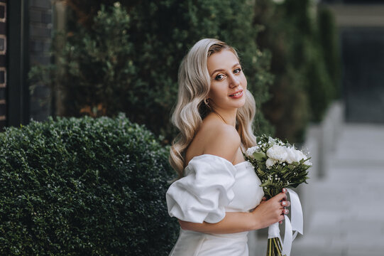 A beautiful, young blonde bride in a white dress with a bouquet of peonies sits near the green thuja, cypress. Wedding photography, portrait.